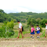 Four young siblings wearing school uniforms walk across a crop field