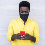 Black man wearing a face mask and leaning against a wall looking at a mobile phone